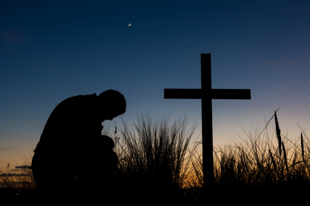 Homem rezando em frente a uma cruz, enquanto o dia escurece. 
