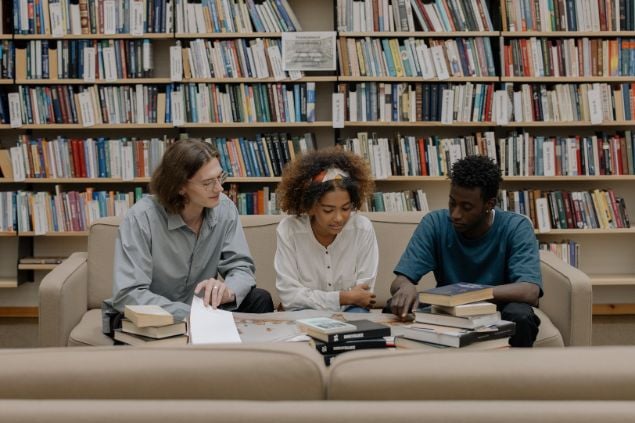 Imagem de três amigos em uma biblioteca conversando e lendo livros, como se estivessem estudando