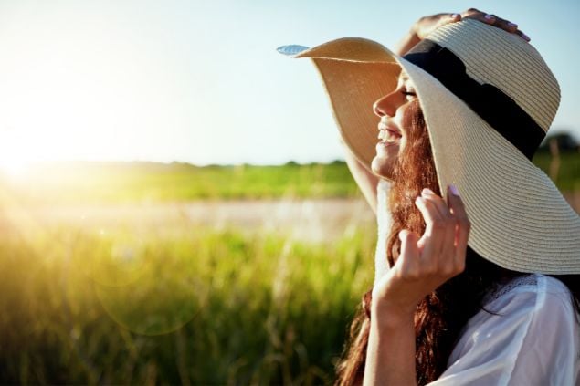 Imagem de uma mulher em um campo sorrindo, o céu está azul e o sol bate em seu rosto
