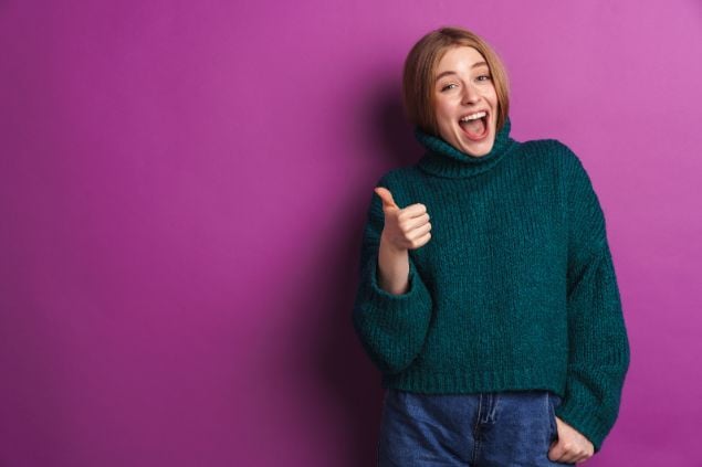 Imagem de uma mulher fazendo sinal de positivo com o dedo e sorrindo em um fundo roxo
