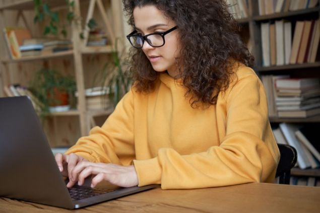 Mulher de óculos e com roupa amarela na frente de seu laptop trabalhando