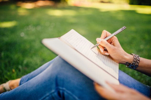 Imagem de uma mulher sentada em um gramado e escrevendo em um caderno de anotações