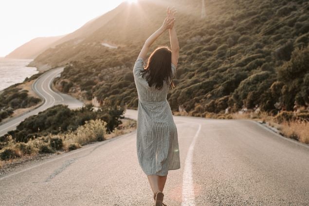 Foto de uma mulher em uma estrada, de costas e com os braços erguidos para cima