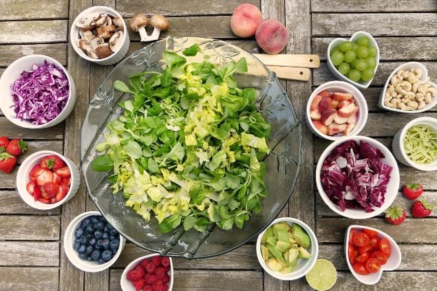 Imagem de uma travessa com salada verde e em volta outras vasilhas menores com legumes e frutas na mesa