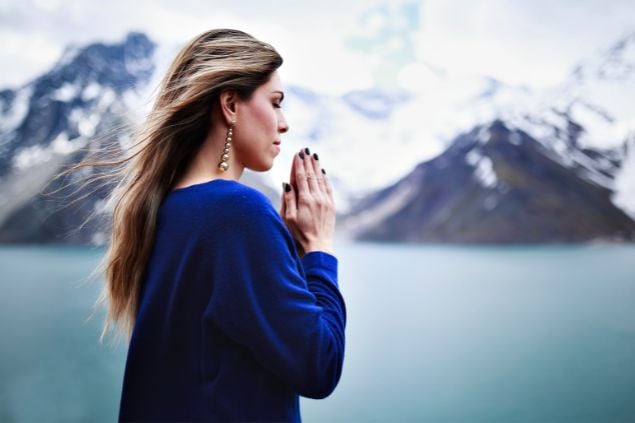 Imagem de uma mulher em uma paisagem de montanhas e lago ao longe, com as mãos juntas e olhos fechados como se estivesse fazendo uma oração