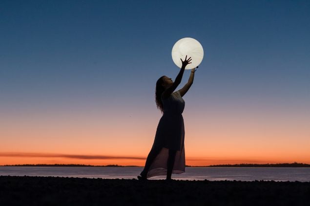 Imagem de uma mulher perto de um lago ao final da tarde com as mãos estendidas para a lua, como se estivesse segurando ela