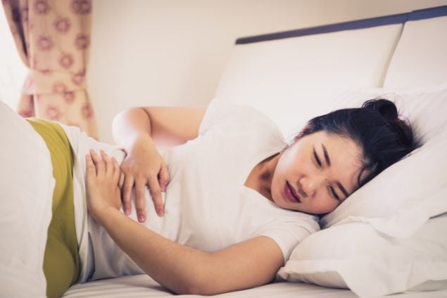 Imagem de uma mulher deitada na cama com as mãos no estômago como se estivesse com dor