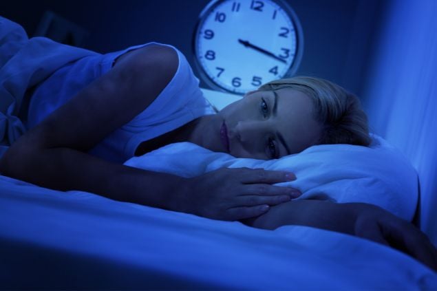 Imagem de uma mulher deitada na cama com os olhos abertos como se tivesse perdido o sono e o relógio atrás marcando 3:15 da madrugada