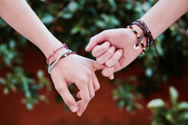 Imagem de duas mãos entrelaçadas pelos dedos mindinhos, os braços de ambos tem pulseiras