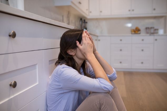 Imagem de uma mulher sentada no chão da cozinha com as mãos no rosto como se estivesse preocupada e triste