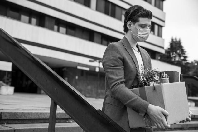 Imagem em preto e branco de um homem de máscara com uma caixa na mão e suas coisas dentro, dando a entender que está saindo do emprego