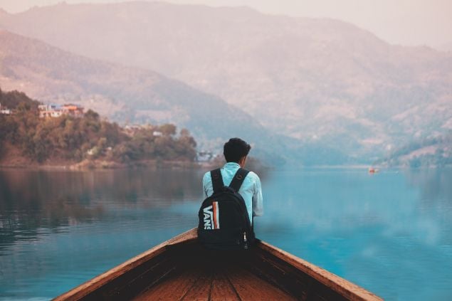 Imagem de um homem na ponta de um barco no lago, e uma paisagem de montanha ao seu redor
