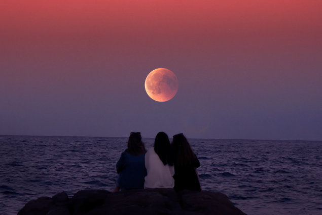 Três meninas sentadas sob uma pedra assistindo à Lua Cheia durante o pôr-do-sol