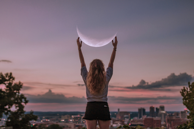 Mulher de costas, com os braços esticados tentando alcançar a lua no céu