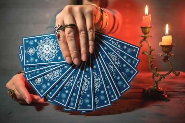 Imagem de uma mão feminina segurando várias cartas de tarot. Ao lado da mesa, uma castiçal com duas velas acesas.