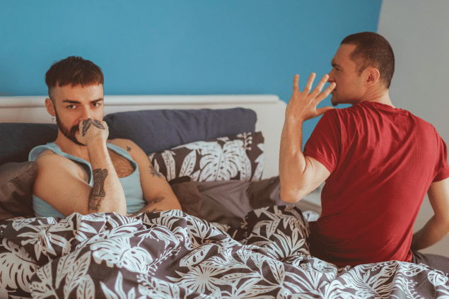 Homem agitando discutindo com seu namorado em quarto. Ambos estão sentados na cama. 