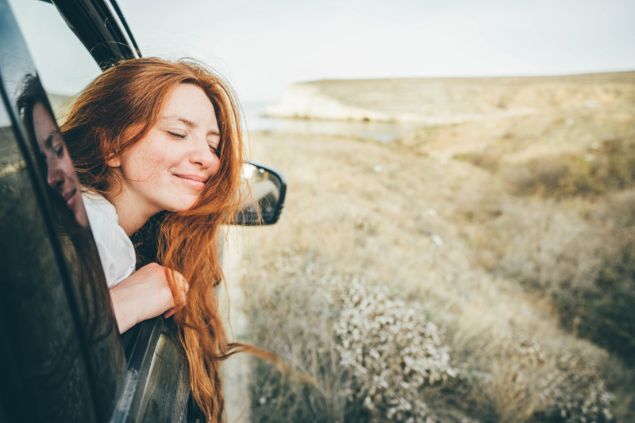 Mulher sorridente, com os olhos fechados, colocando a cabeça para fora da janela de um carro em movimento.