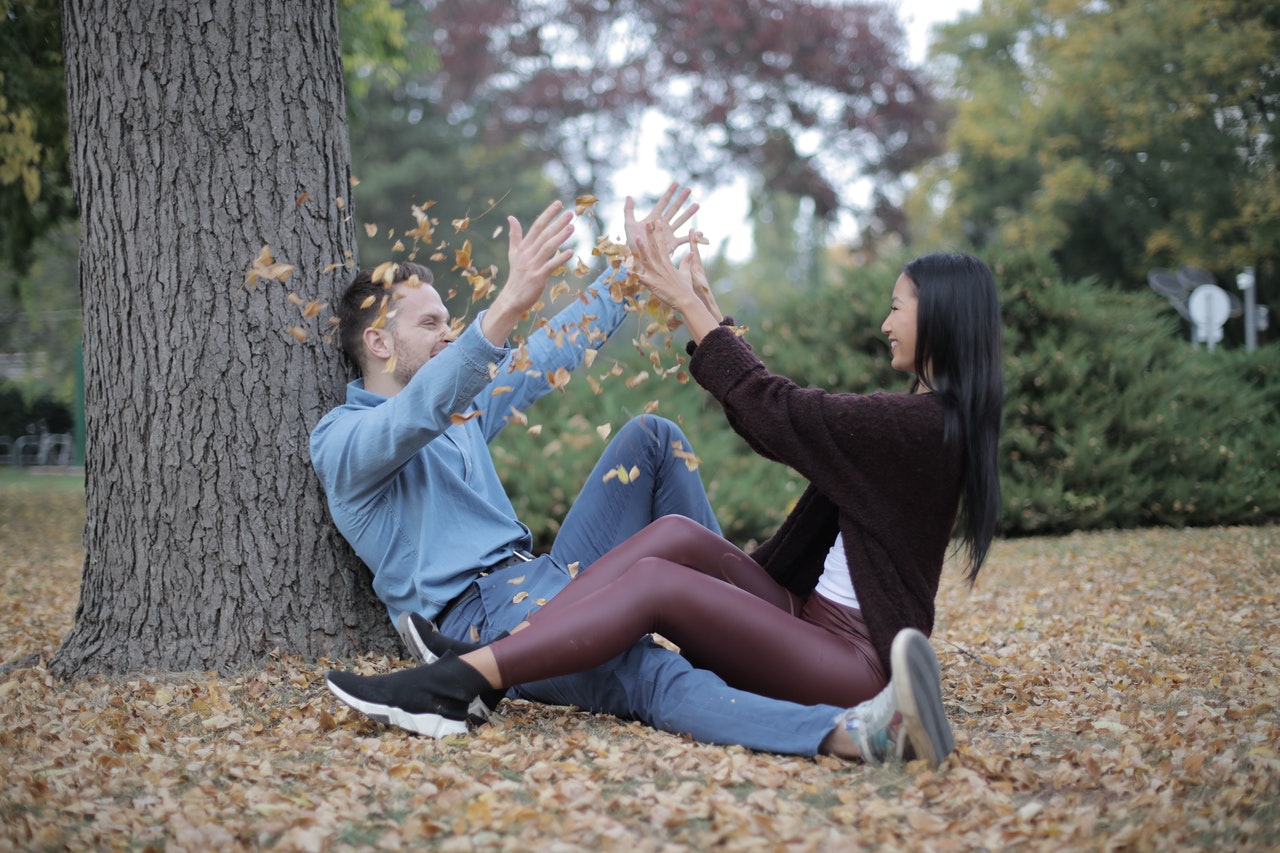 Homem e mulher sentados em parque, jogando folhas secas um no outro.