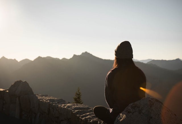 Mulher de costas sentada numa pedra observando montanhas.