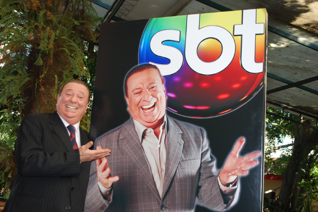 Raul Gil ao lado de um cartaz com seu rosto e com o logo do SBT