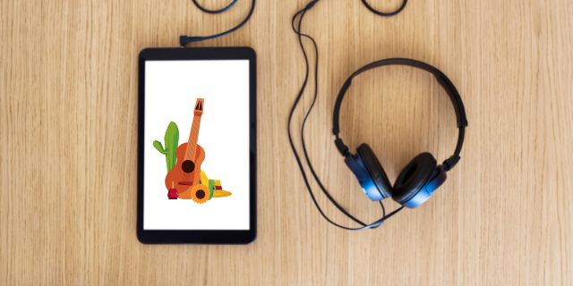 Fones conectados a tablet - ambos estão sobre superfície lisa e clara de madeira. Na tela do tablet, há elementos remetentes à comemoração de festa junina: violão, cacto, um girassol, chapéu de palha, milho e uma maçã do amor.