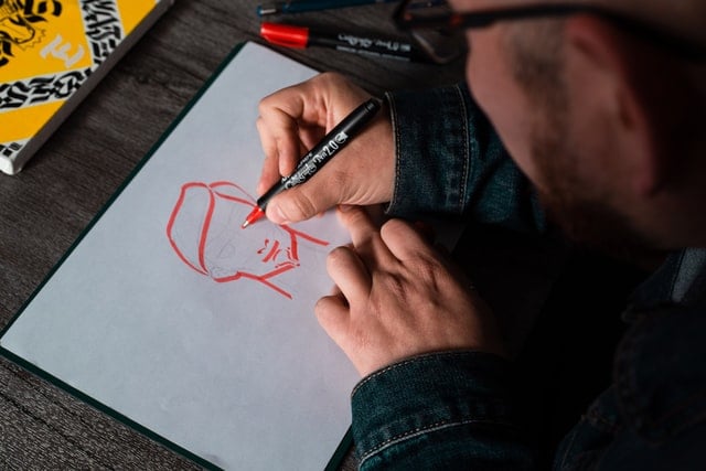 Homem branco desenhando uma pessoa com caneta vermelha.