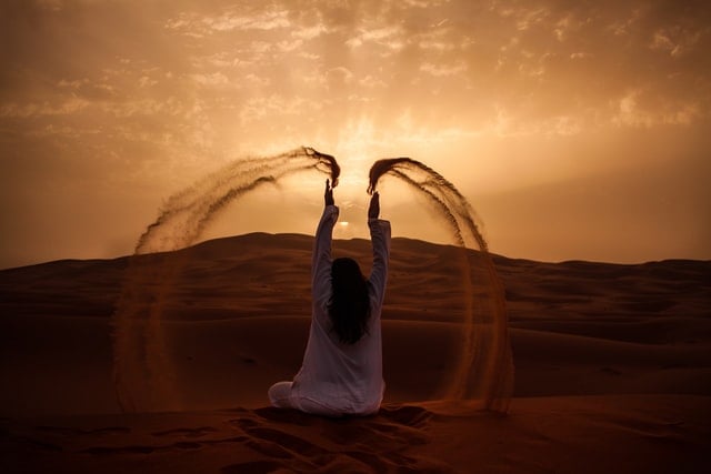 Silhueta de mulher sentada de costas num deserto, jogando areia para cima.