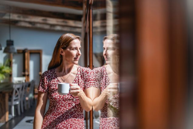 Imagem de uma mulher na frente de uma janela tomando café na caneca e refletindo sobre a vida
