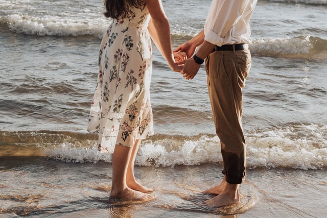Casal de mãos dadas na praia, com os pés tocando o mar