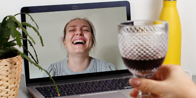 Mulher segura taça de vinho em frente de computador. Na tela, há outra mulher sorrindo. As duas estão em uma vídeo-chamada.