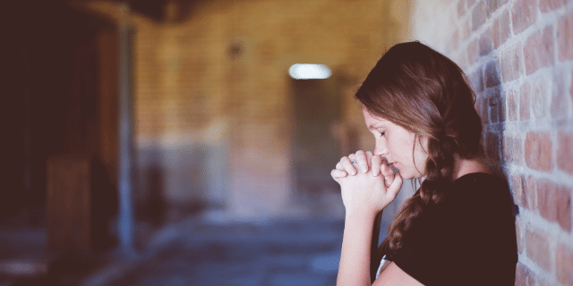 Uma mulher com os olhos fechados rezando encostada na parede. 