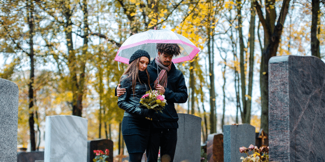 Um casal abraçados de roupa preto debaixo do guarda-chuva dentro de um cemitério olhando para um túmulo. 