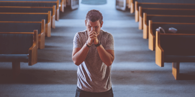 Um homem ajoelhado na igreja rezando de olhos fechados. 