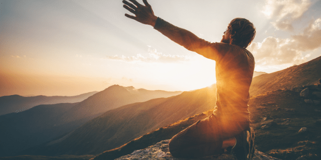Um homem ajoelhado com os braços abertos rezando em meios as montanhas. 