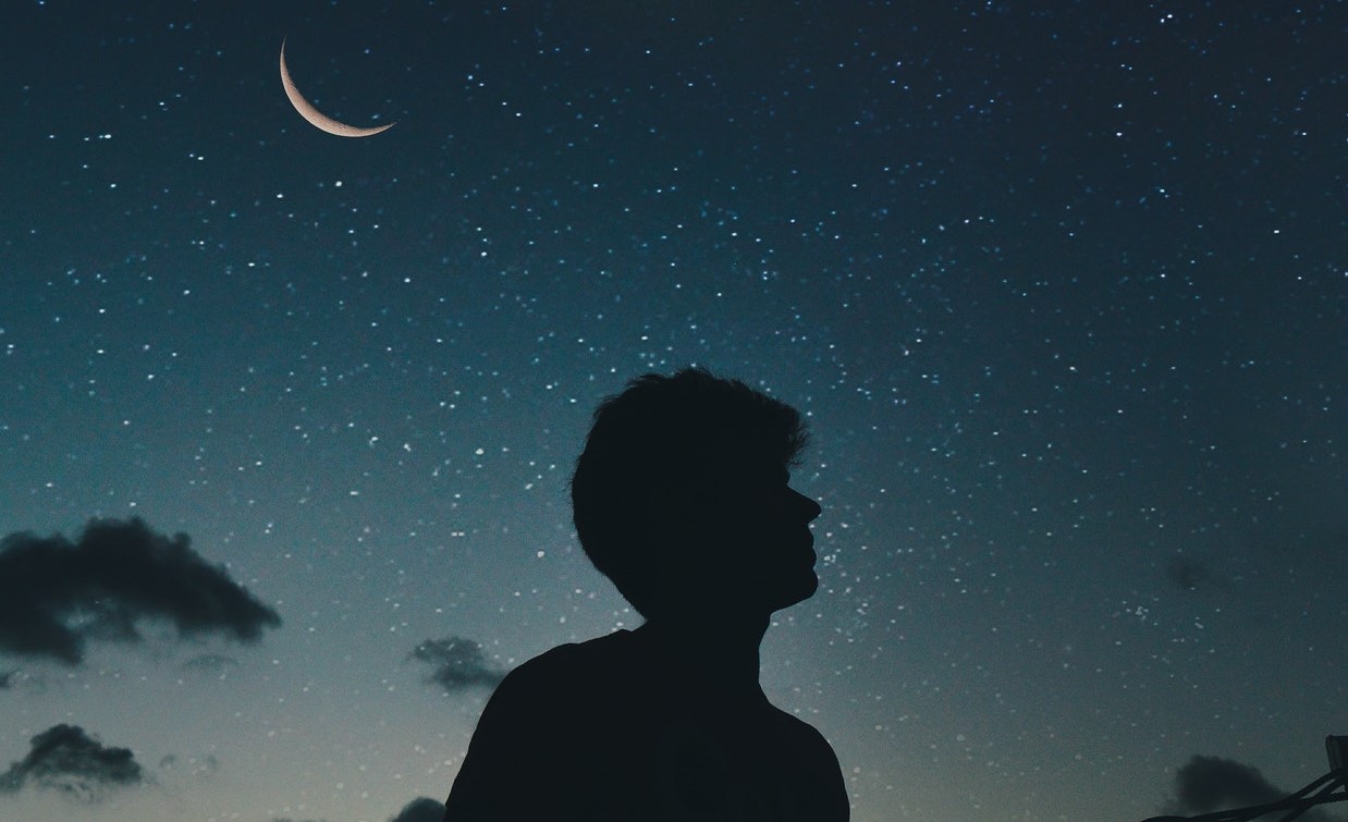 Silhueta de um homem com o céu estrelado de fundo e a lua posicionada à esquerda 