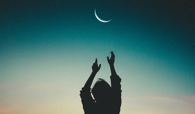 Silhueta de uma pessoa com os braços levantados tentando alcançar a Lua Nova no céu