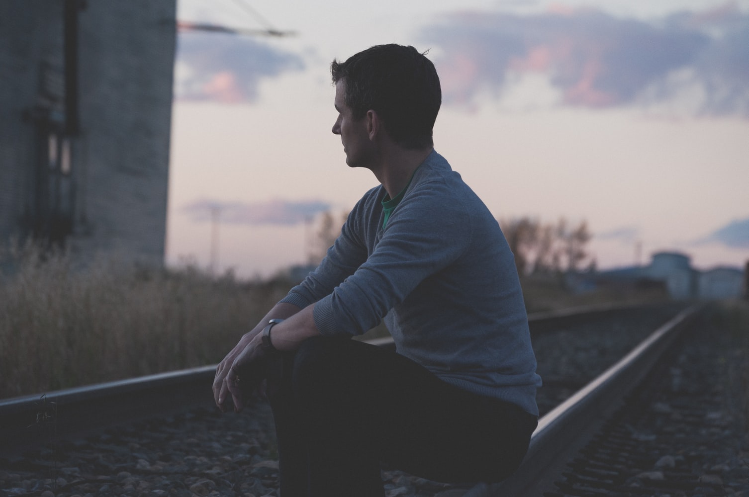 Homem sentado de lado em um trilho de trem no fim da tarde olhando para o horizonte