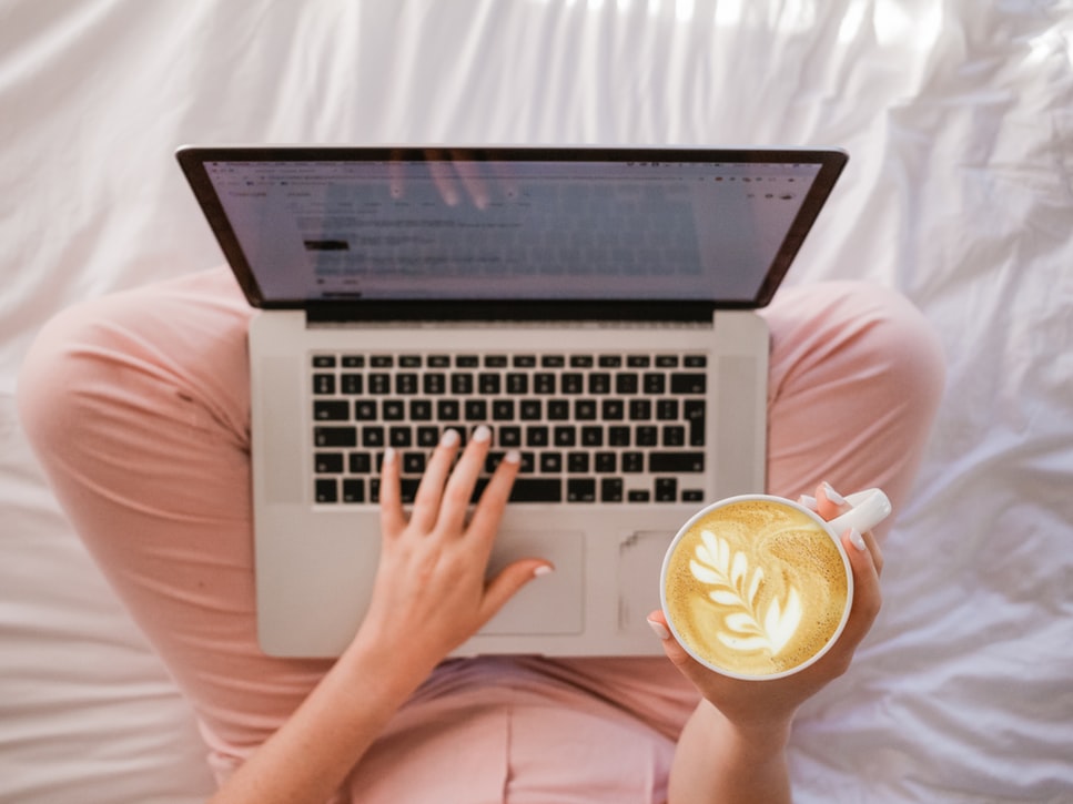 Mulher sentada na cama trabalhando com um computador, enquanto segura uma caneca de café.