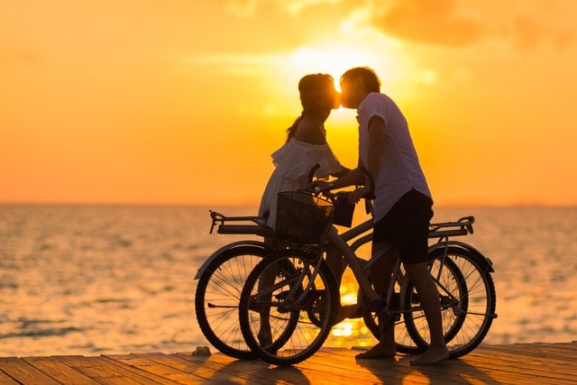Homem e mulher segurando suas bicicletas enquanto se beijam ao pôr do sol.