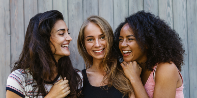 Três mulheres sorriem e olham umas para as outras. 